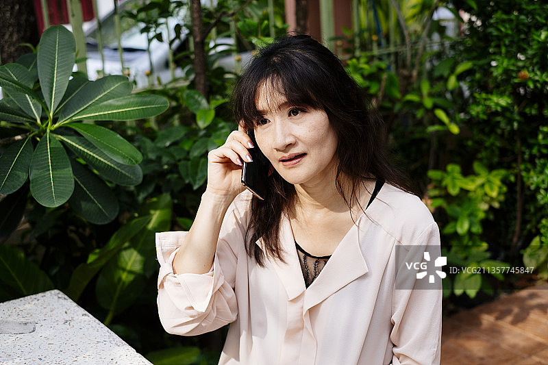 成年黑头发亚洲女人坐在公园的长凳上讲电话。图片素材