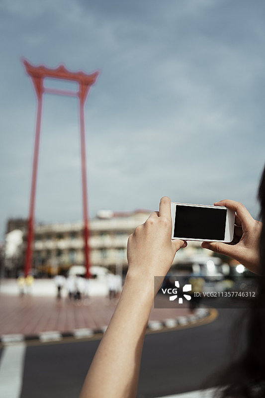 一名泰国女子举起智能手机，为巨大的秋千拍照，这是一张裁剪过的照片。图片素材