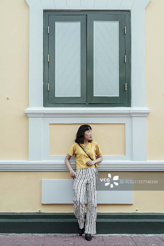 卷发短发的泰国时尚达人，身穿黄色t恤和脱裤，站在淡黄色的建筑前，优雅的姿势就像一个超级名模。图片素材