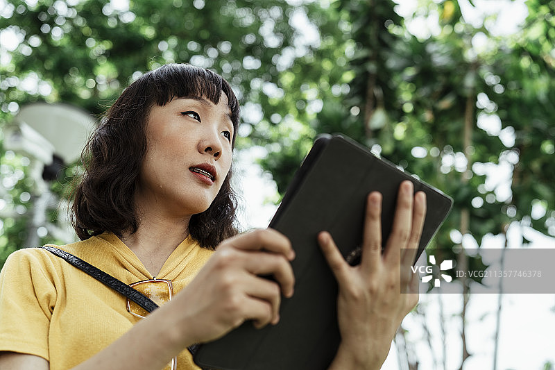 卷发短发的泰国时尚达人身穿黄色t恤，手拿平板，与散景一起站在绿树成荫的公园里。图片素材