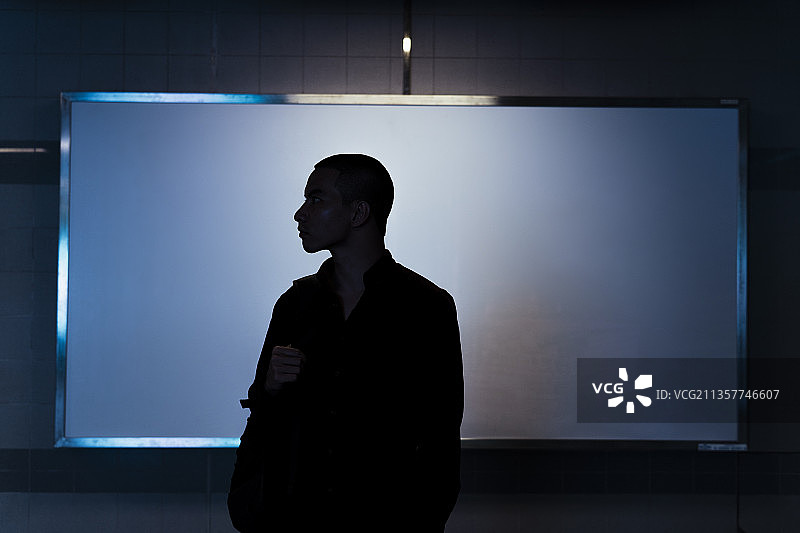 光头男子背着背包，站在白板前一盏灯下，剪影。图片素材