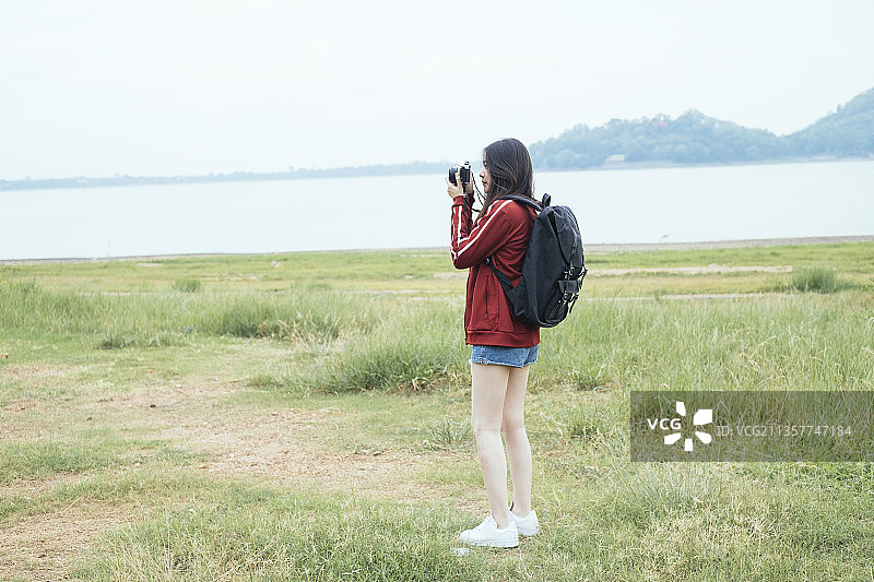年轻的亚洲泰国人，深色头发的旅行者女人穿着红色夹克在春武里湖拿着相机。图片素材