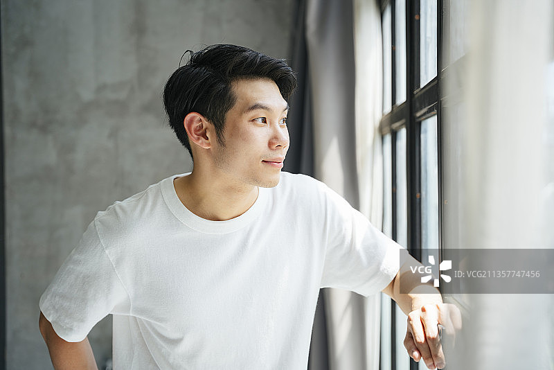 一个穿着白衬衫的亚洲年轻人在家里看着窗外。图片素材