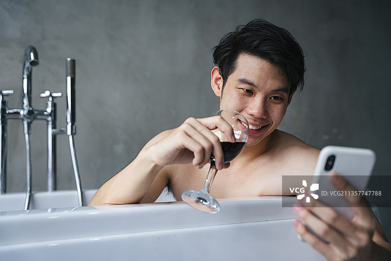 亚洲年轻人在浴缸里喝酒，用智能手机。图片素材