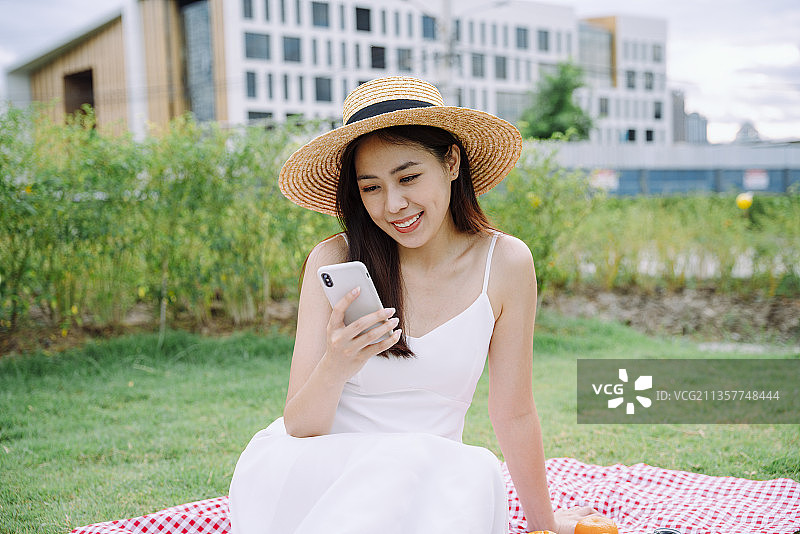 美丽的亚洲旅行者女人穿着白色的裙子和帽子在公园户外使用智能手机。图片素材