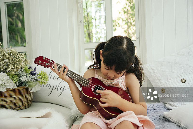 亚洲童女用木制经典尤克里里在家里的沙发上。图片素材