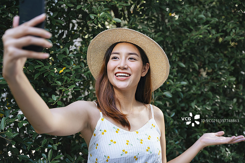 美丽的亚洲女性戴着帽子，身穿白色连衣裙，在公园里自拍或用智能手机拍旅游视频。图片素材