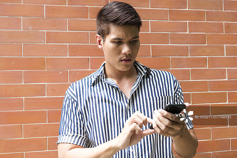 一名亚洲男子站在砖墙背景下，用智能手机做网上银行业务。图片素材