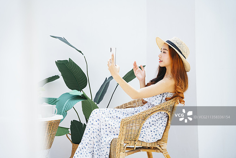 侧视图-美丽的亚洲旅游妇女戴着帽子使用智能手机在椅子上。图片素材