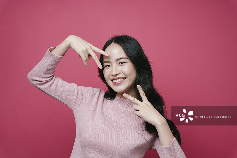 年轻亚洲妇女的肖像显示胜利的姿势与两个手指孤立在粉红色的背景。图片素材