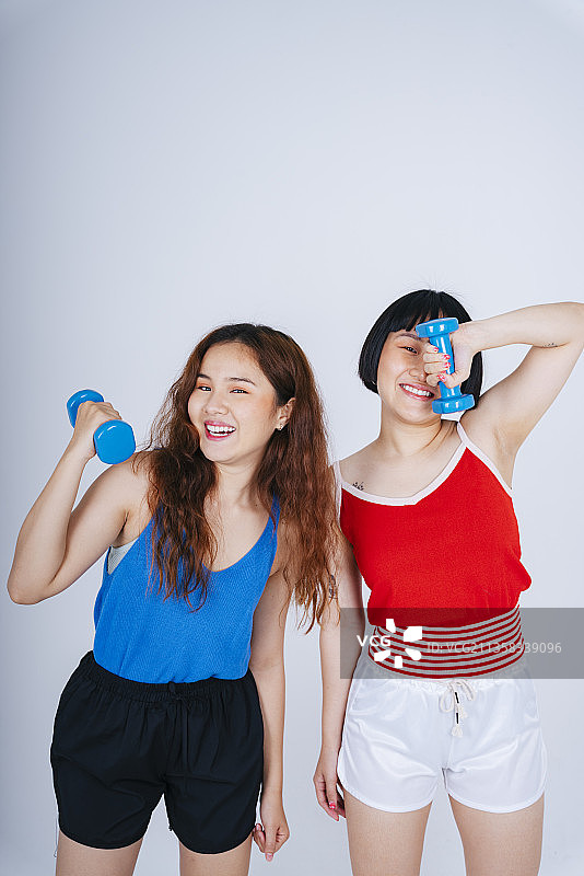 两个年轻的亚洲妇女在蓝色和红色的衬衫拿哑铃孤立在白色的背景。图片素材