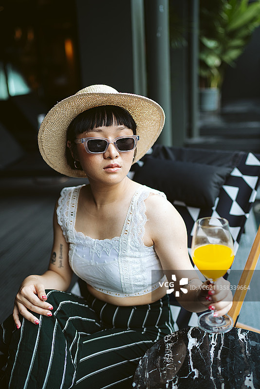 年轻的亚洲妇女戴着太阳镜和帽子在酒店喝橙汁。图片素材