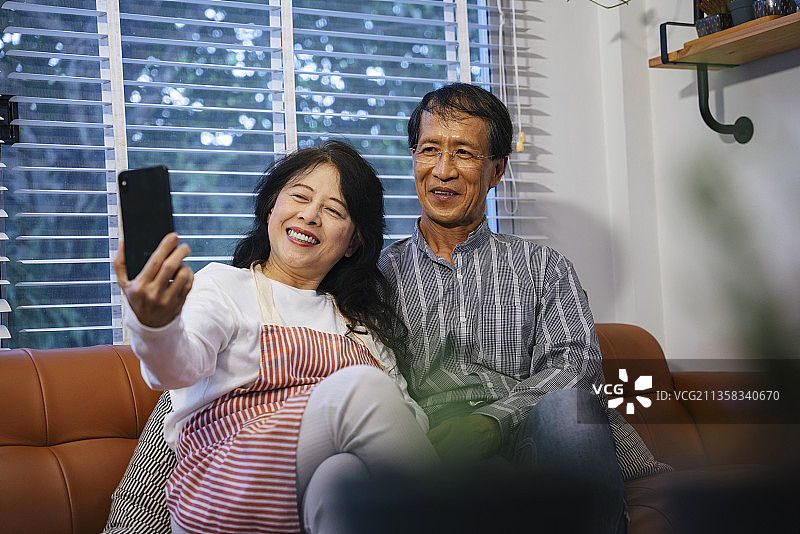 亚洲老年夫妇一起在家里沙发上用智能手机自拍。图片素材