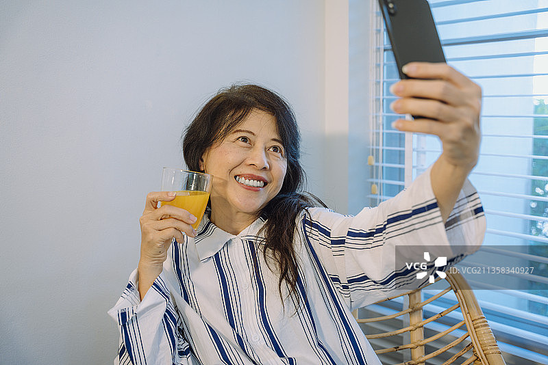亚洲高级女性拿着橙汁用智能手机自拍。图片素材