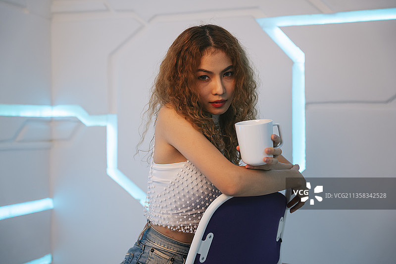 时髦的亚洲卷发女人的肖像坐在椅子上持有陶瓷咖啡杯在现代的房间。图片素材