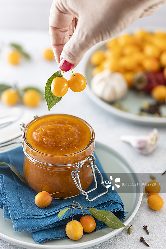 女人手持黄色樱桃李，在酱料酸辣酱玻璃罐上方图片素材