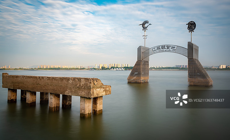 河中间矗立着长江中的江豚观赏地标志，金属拱门，两边柱子上江豚的金属雕像，一旁有一条断裂的堤坝图片素材