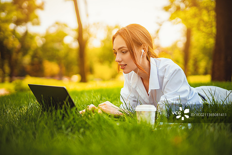 年轻红发女学生躺在草地上使用笔记本电脑和耳机图片素材