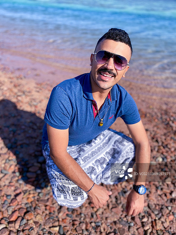微笑的年轻人戴着太阳镜坐在埃及达哈布海滩上的肖像图片素材