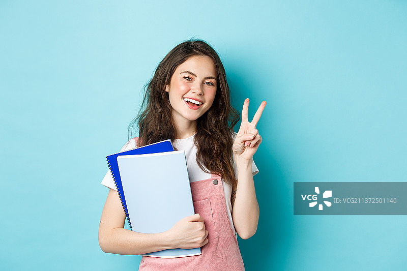 拿着笔记本，面带微笑，做出V字手势的漂亮女学生图片素材