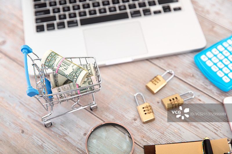 笔记本电脑和手机及载有美元钞票的购物车模型围绕着三把密码锁图片素材