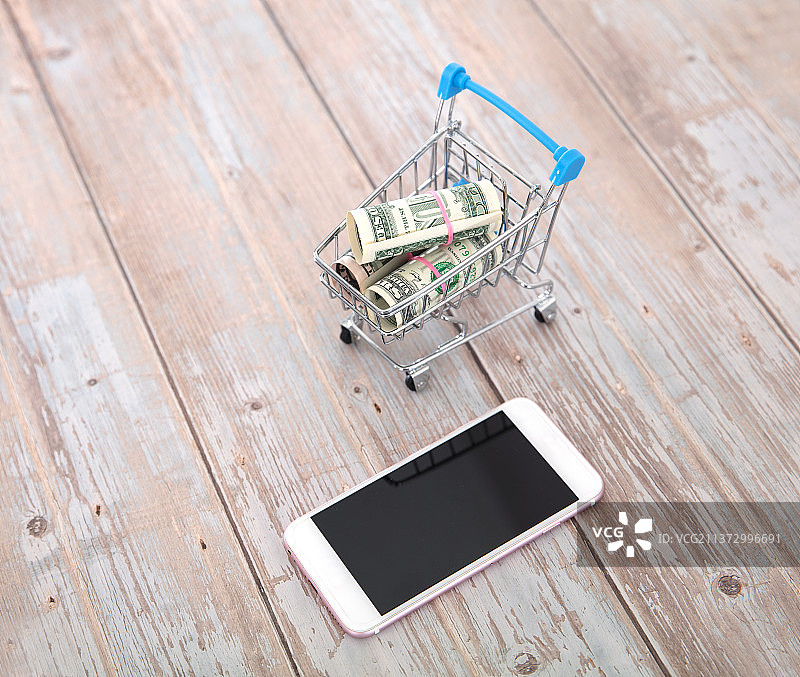 桌子上满载美元钞票的购物车和一部智能手机图片素材