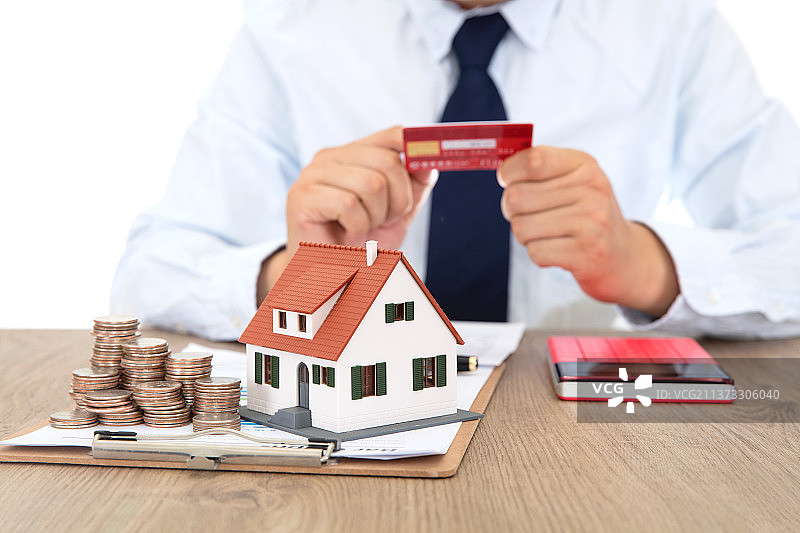 桌子上放置着小房子模型和美元硬币及看着银行卡的商务男性坐在桌旁图片素材