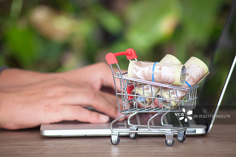 装满加元钞票的购物车模型在双手正打字的笔记本电脑旁边图片素材