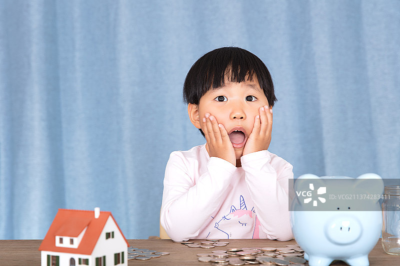 在一堆硬币和一个存钱罐及一个房子模型前做鬼脸的孩子图片素材
