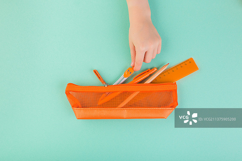 孩子的手拿着橙色袋子里的所有铅笔、钢笔、剪刀，背景为蓝绿色图片素材