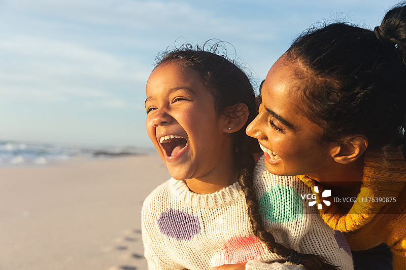 年轻的混血女子和女儿在日落时分在海滩上对着天空欢笑图片素材
