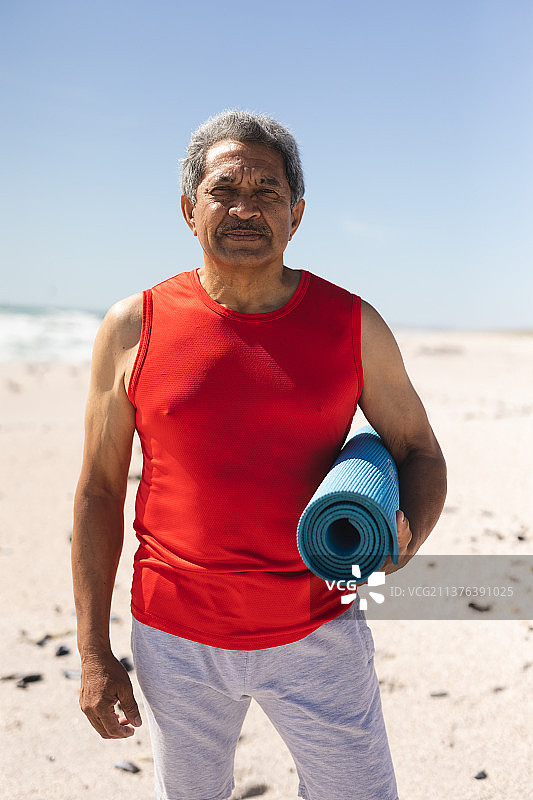 自信的混血老年男人拿着卷起的瑜伽垫在海滩上，蓝色天空图片素材