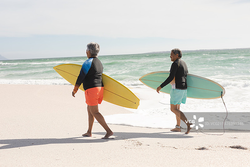 多种族老年夫妇在阳光明媚的海滩上扛着冲浪板走在岸边图片素材