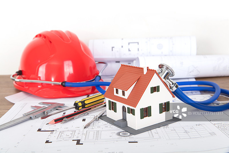 工程图纸和图纸上一个带着听诊器正给一个小房子模型听诊的安全帽和其它相关工具图片素材