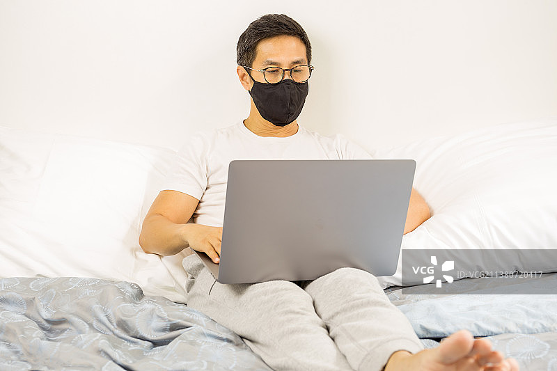 疫情期间，戴口罩男子在家中床上用笔记本电脑工作图片素材