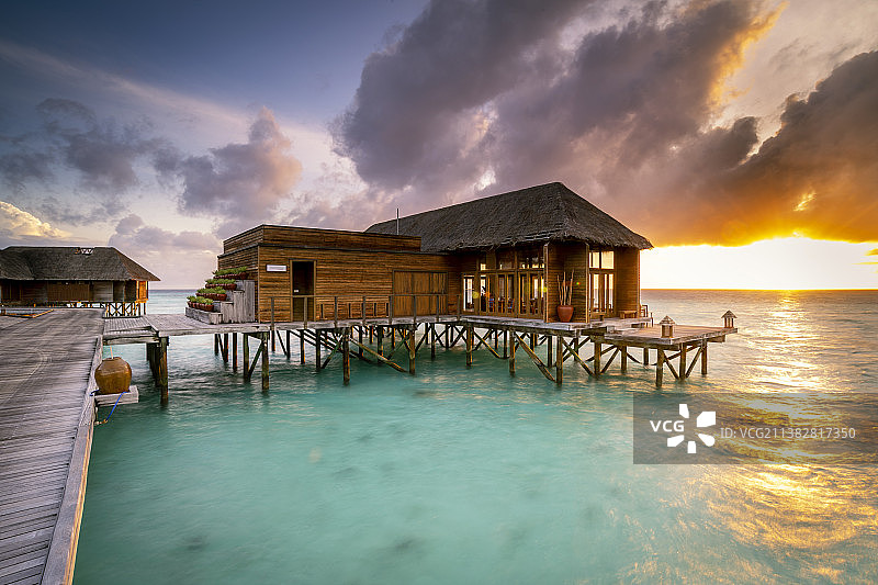 日出时分热带珊瑚礁，蓝色天空朝霞满天，栈桥和一排排水上度假木屋排开，温暖太阳跳出海面图片素材