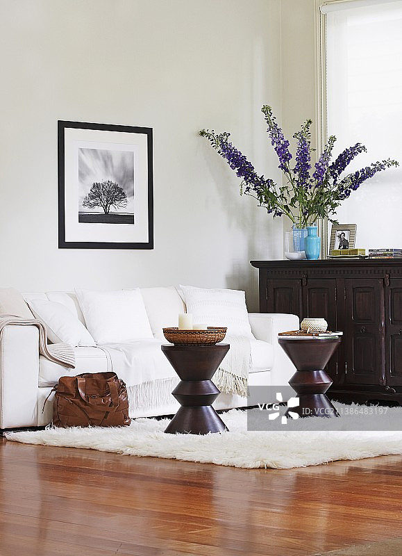优雅的起居区，配有光面拼花地板、靠墙的白色沙发、深色鼓形边桌以及深色木柜上的一束紫色鲜花图片素材