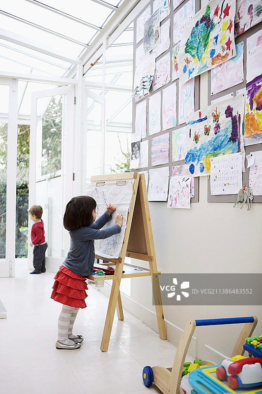 在画架上绘画的小女孩，儿童画作贴在带有玻璃天花板的白色内墙上图片素材