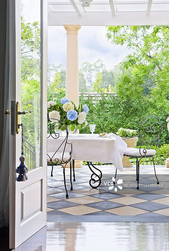 透过敞开的门看到的露台景色，露台铺有棋盘格瓷砖，锻铁椅子摆放在白色桌子旁图片素材