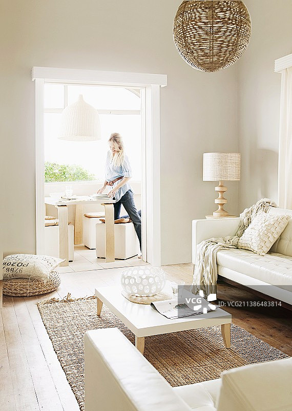 白色矮咖啡桌置于剑麻地毯上，浅色沙发套装正对着敞开的门口，可见一名妇女正在摆放桌子图片素材