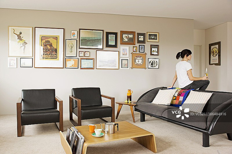 画廊墙前的浅色木质现代咖啡桌和黑色皮革扶手椅；女子坐在灰色沙发后背上图片素材