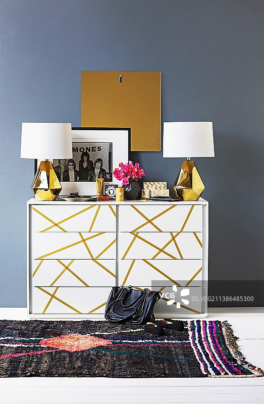 蓝灰色墙壁前金色条纹白色抽屉柜上的黄铜底座白色灯罩台灯图片素材