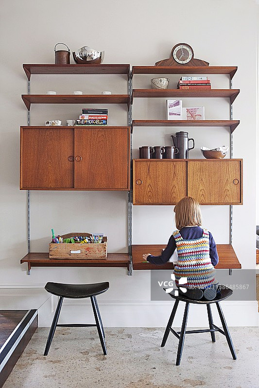 小女孩坐在木凳上，面前是复古风格的模块化置物架，带有隔间、柜子和壁挂式书桌图片素材