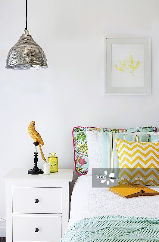 白色床头柜旁边的带各种枕头的床与鹦鹉摆件图片素材