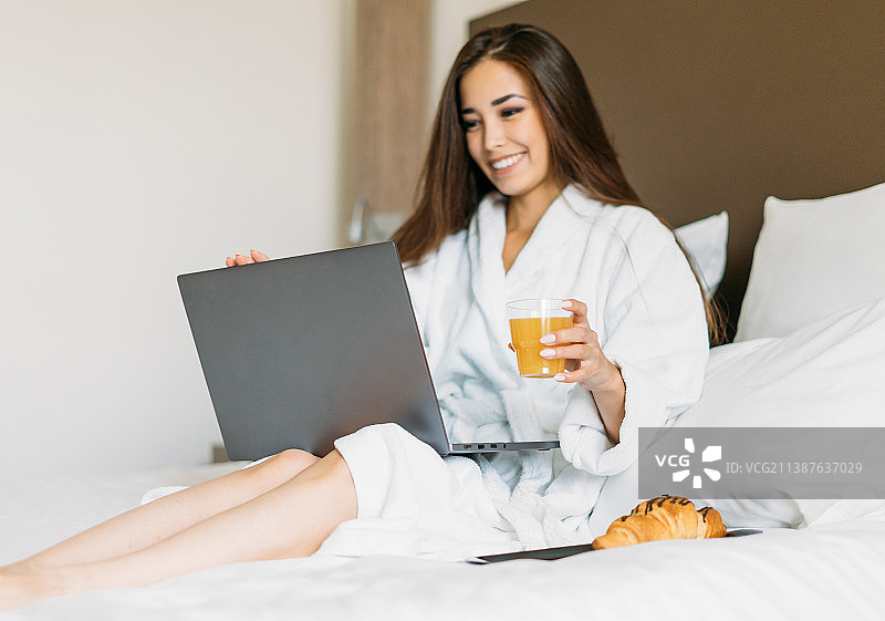 身穿白色长袍的年轻亚洲女孩在酒店房间的床上使用笔记本电脑吃早餐图片素材