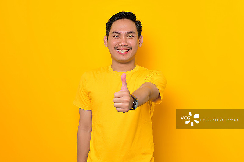 微笑的亚洲年轻男子在黄色背景上竖起大拇指并做出赞许的手势，印度尼西亚图片素材