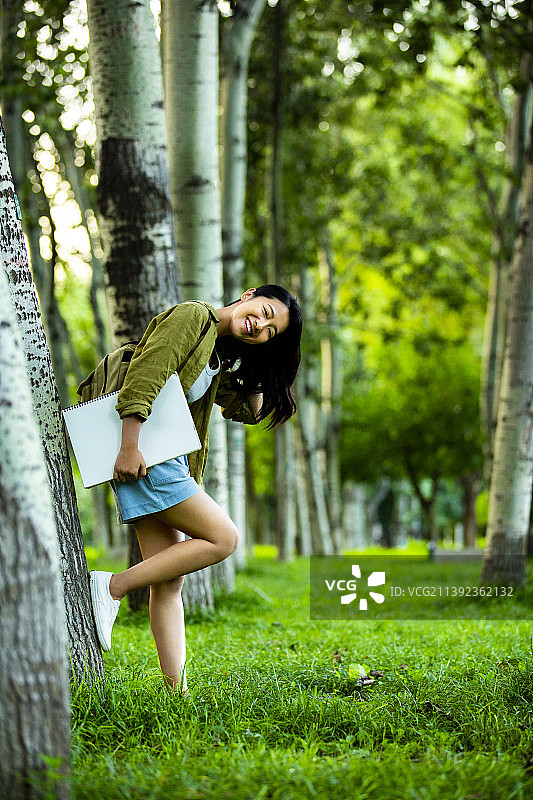 青年女子抱着速写本，倚靠树干休息，在公园寻找灵感，准备画画图片素材