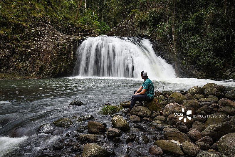男子坐在澳大利亚昆士兰森林瀑布旁的岩石上的侧视图图片素材