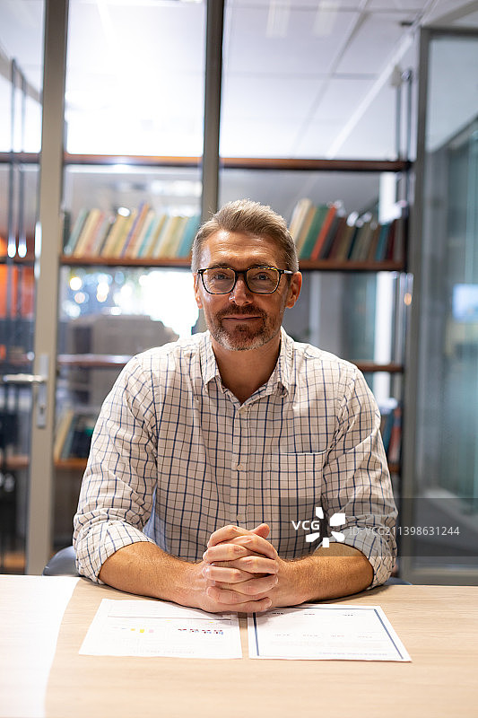 微笑自信的白人男商人坐在工作场所的办公桌前，双手合十的肖像图片素材