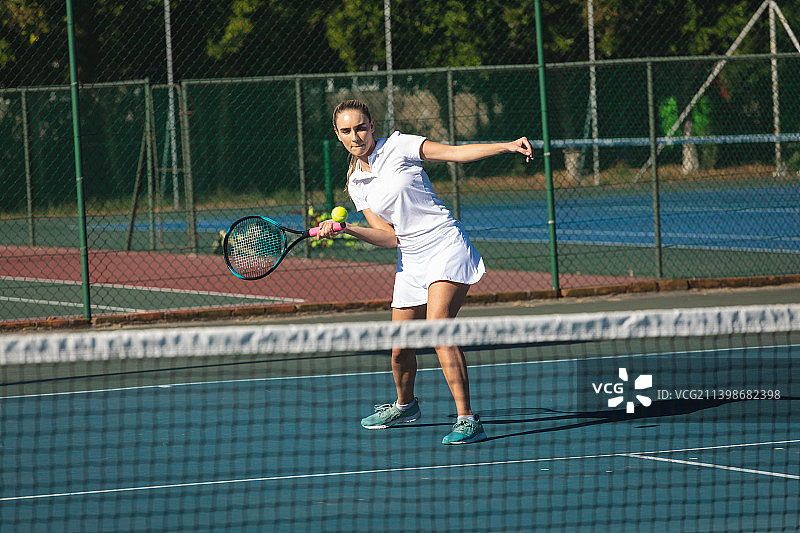 年轻白人女选手在阳光明媚的网球场上用球拍击球全身照图片素材